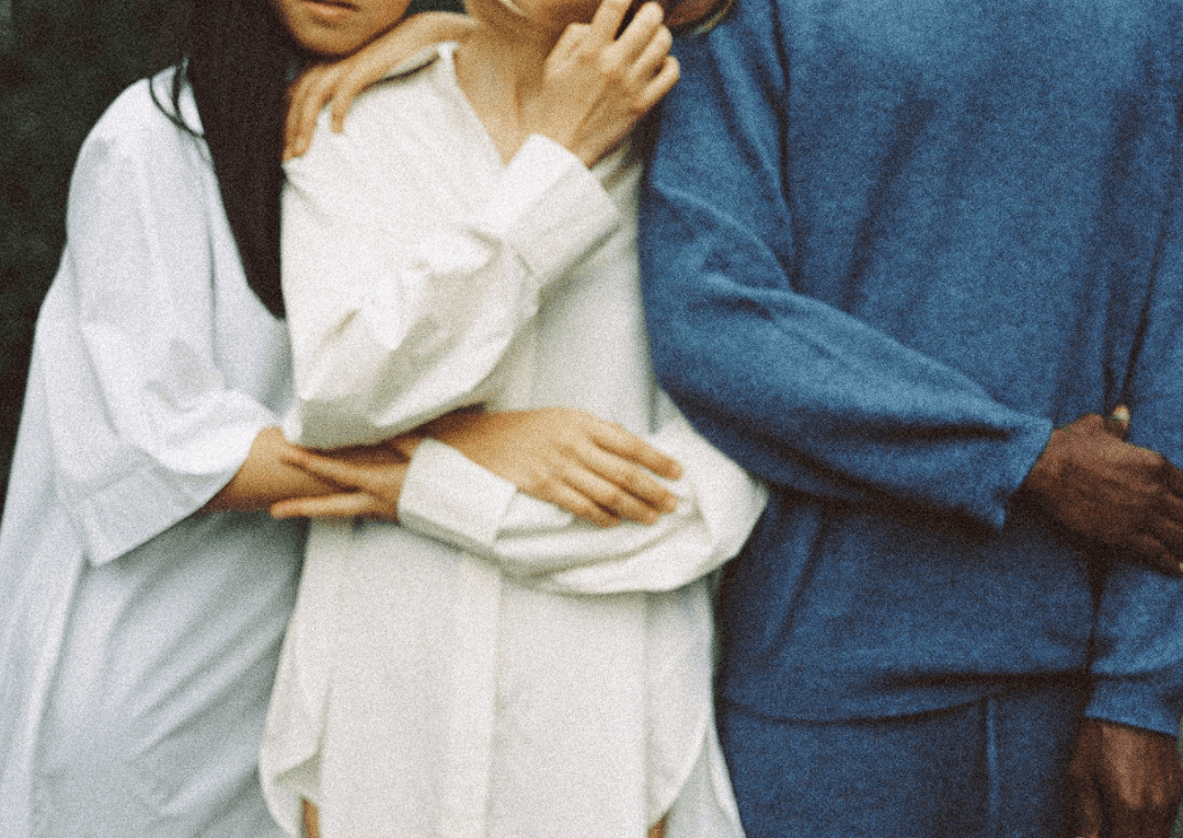 Three people stand next to one another, arms crossed and leaning on each other. The two women on the left are wearing white, and the man on the right is wearing blue.