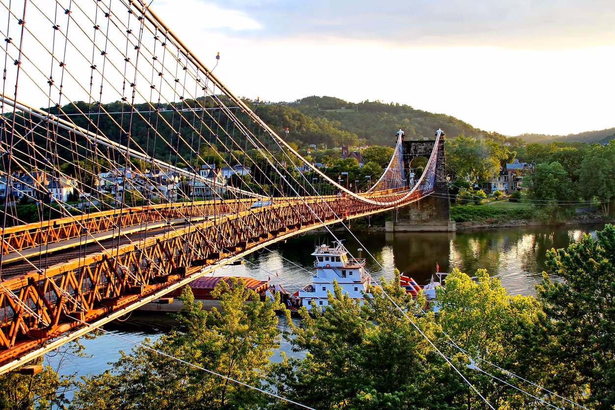 Suspension bridge over a river with a boat passing underneath, surrounded by green trees and houses, with hills in the background.