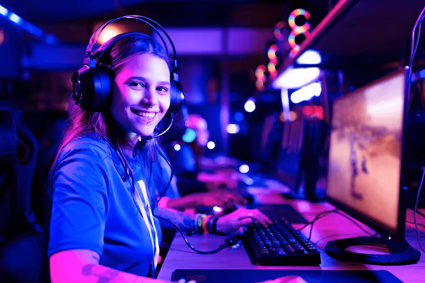 Smiling young woman wearing a headset and gaming at a computer in a blue and purple neon-lit environment.
