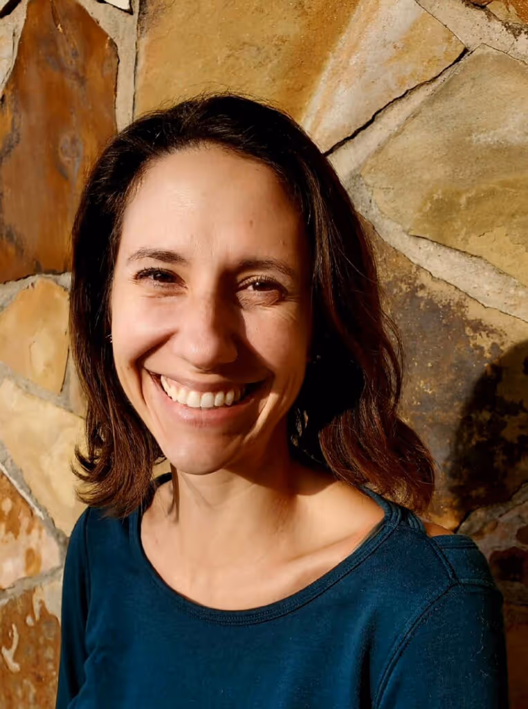 Smiling woman with shoulder-length dark brown hair wearing a blue shirt standing in front of a stone wall.