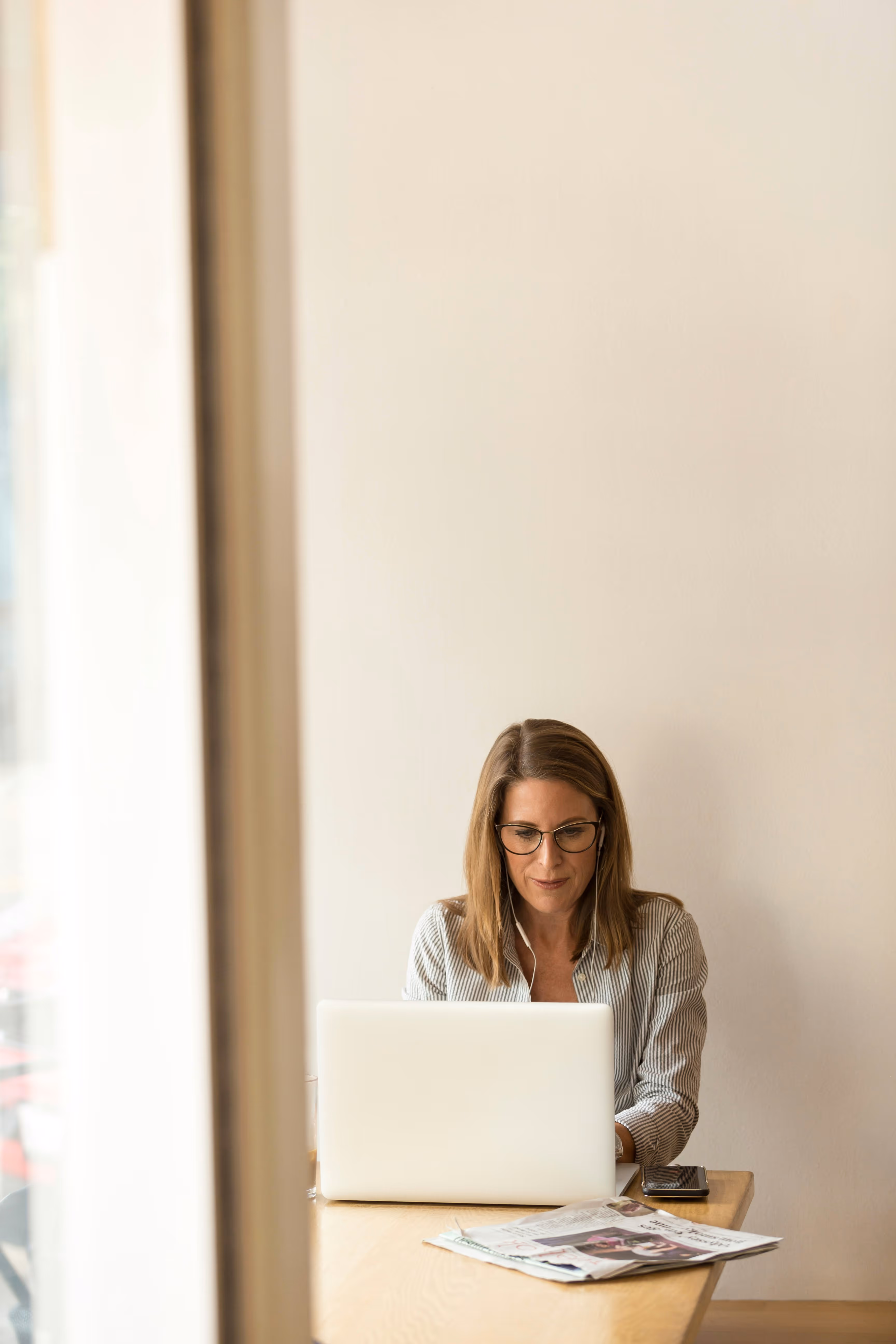 Woman wearing glasses and earphones working on a laptop at a wooden table with newspapers and a phone beside her.
