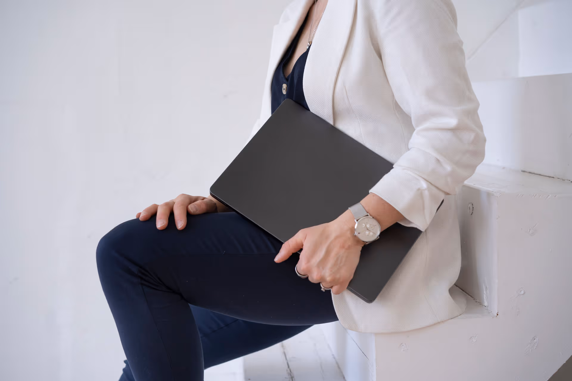 Person sitzt auf weißen Stufen, hält einen schwarzen Laptop und trägt ein weißes Sakko, dunkle Hose und eine Armbanduhr.