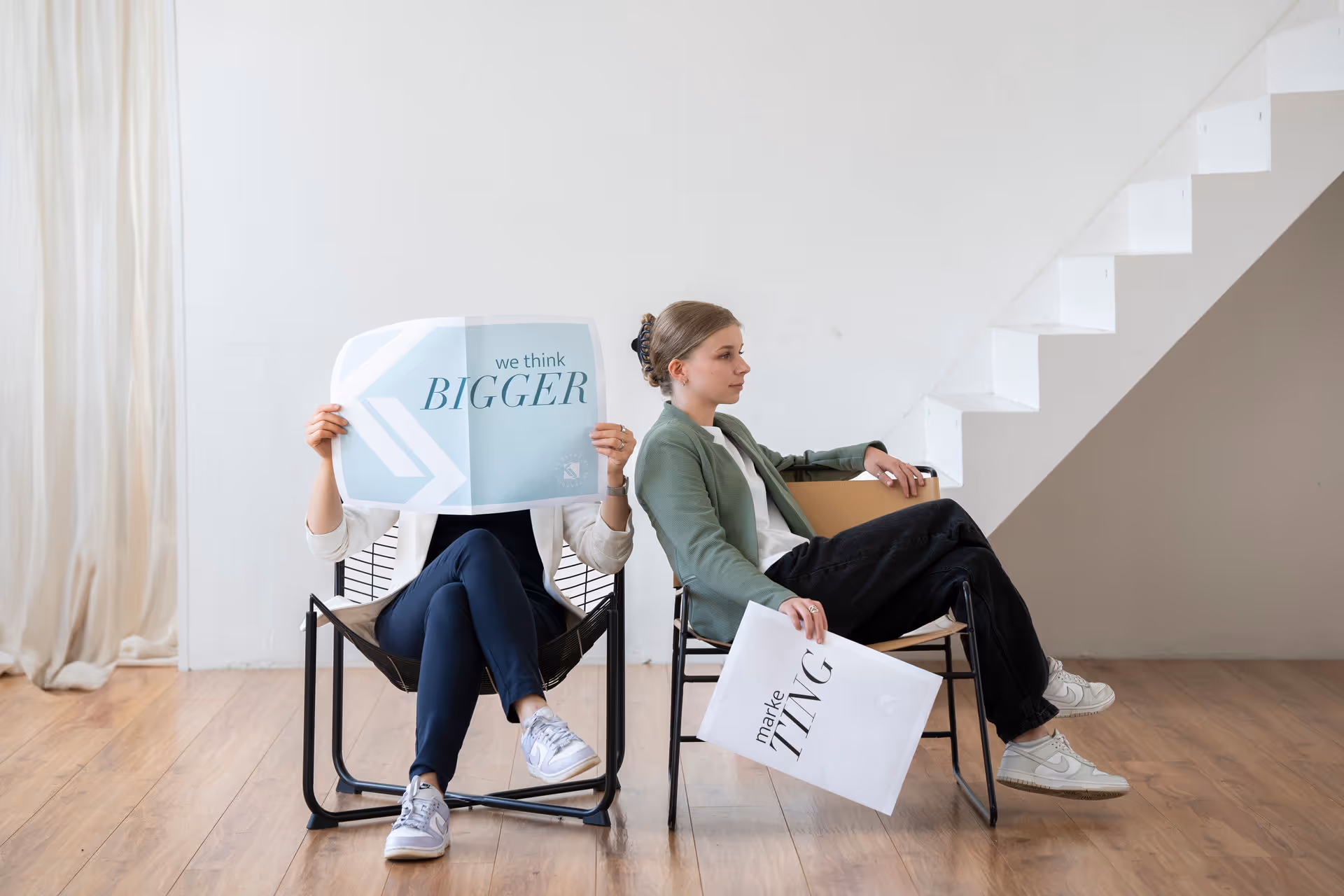 Zwei Frauen sitzen auf Stühlen vor einer weißen Wand, eine hält eine Zeitung mit dem Text 'we think BIGGER', die andere hält ein Blatt mit dem Wort 'marketing'.