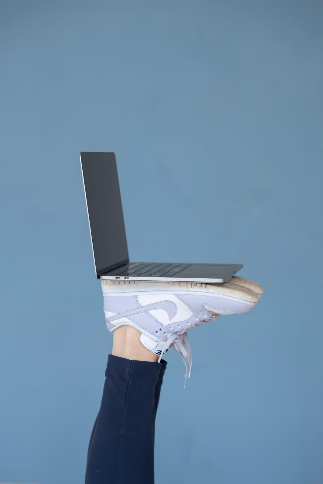 Laptop liegt auf den nach oben gestreckten Füßen einer Person in weißen Sportschuhen und dunkler Hose vor einem blauen Hintergrund.
