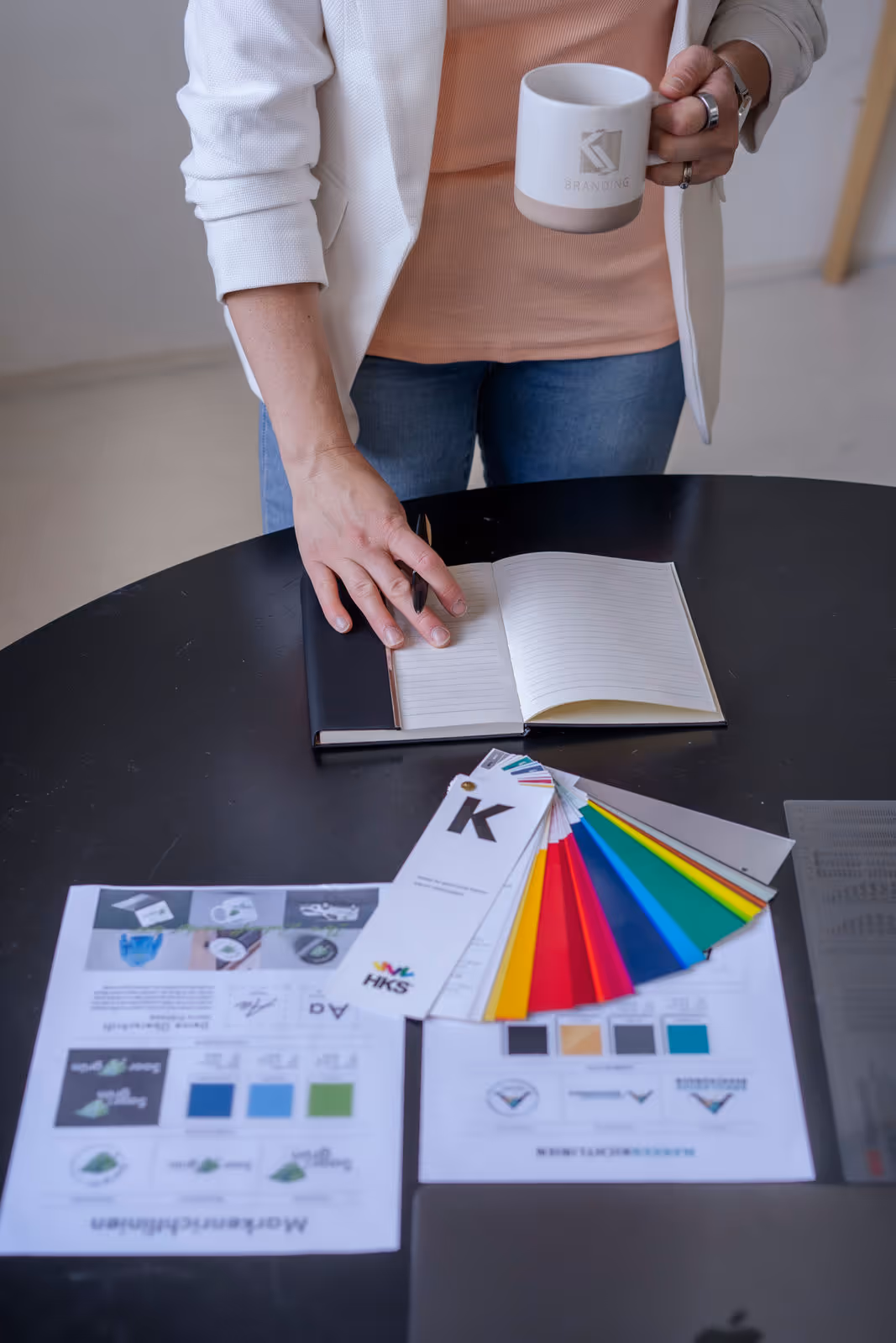 Person steht an einem schwarzen Tisch mit offener Notizbuchseite, einer Farbpalette und gedruckten Markenrichtlinien, während sie eine weiße Tasse mit Branding hält.