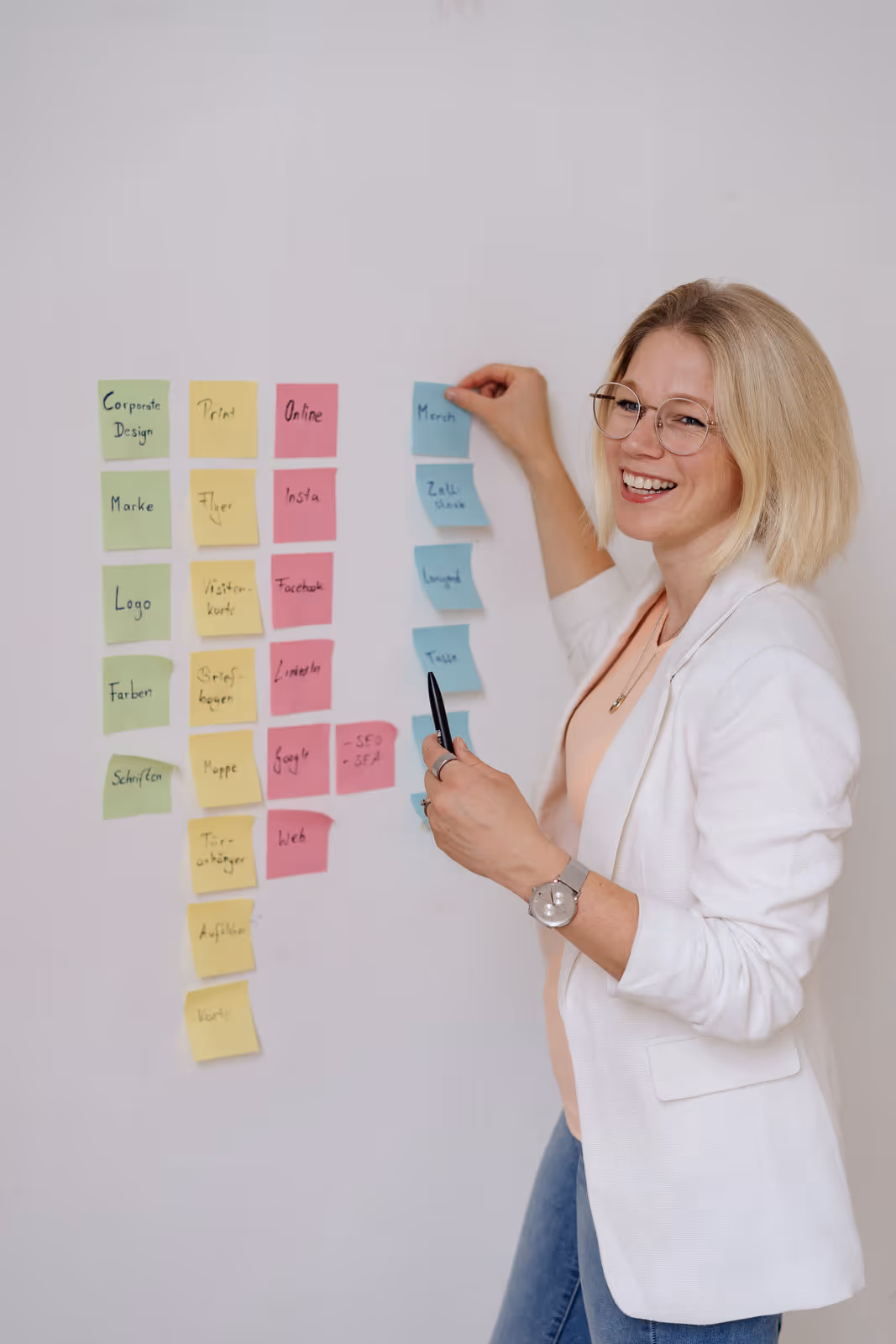 Frau mit Brille und weißem Blazer lächelt, während sie farbige Haftnotizen an eine Wand klebt.