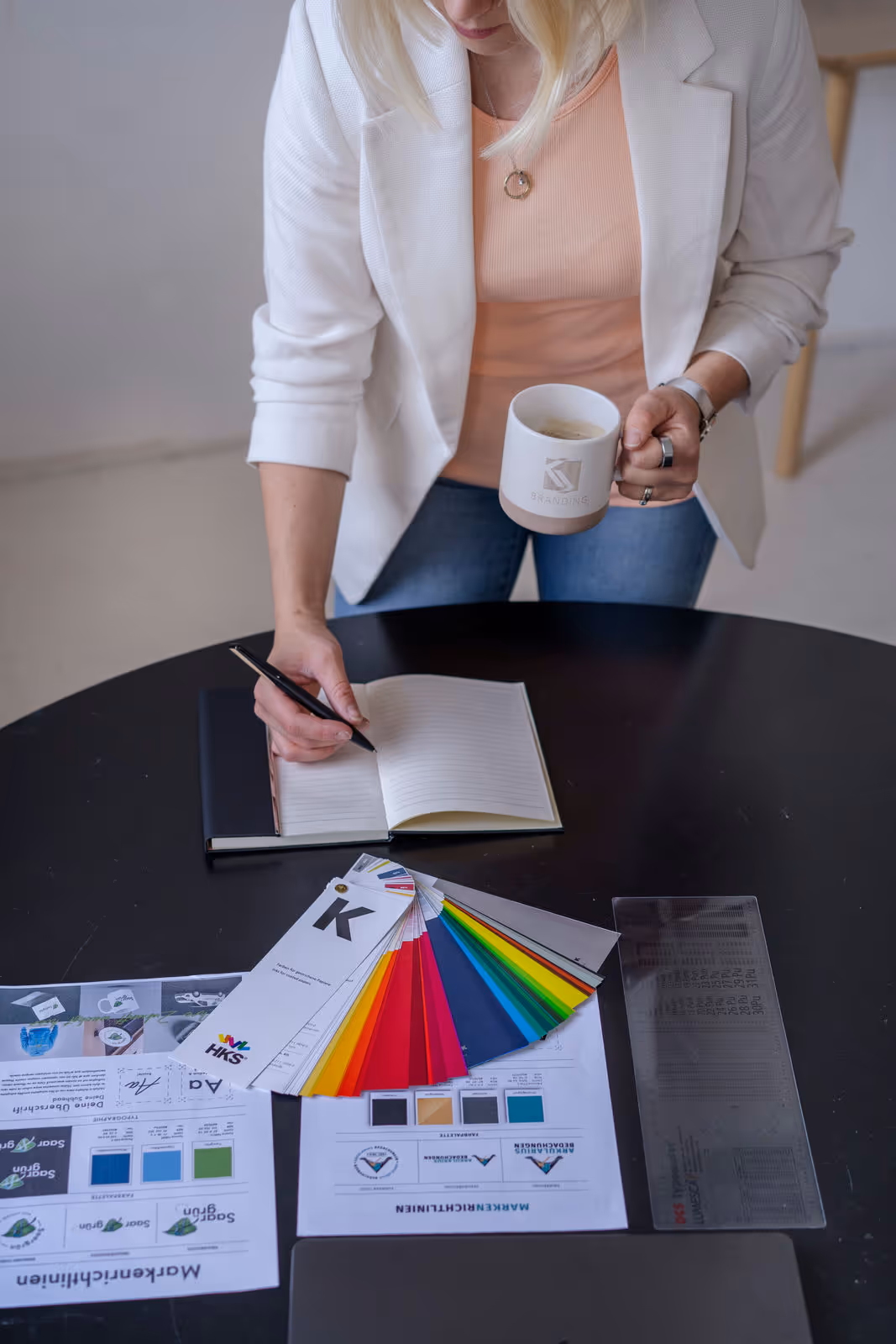 Frau mit weißem Blazer hält eine Kaffeetasse und schreibt in ein Notizbuch auf einem Tisch mit Farbfächern und Marketingunterlagen.