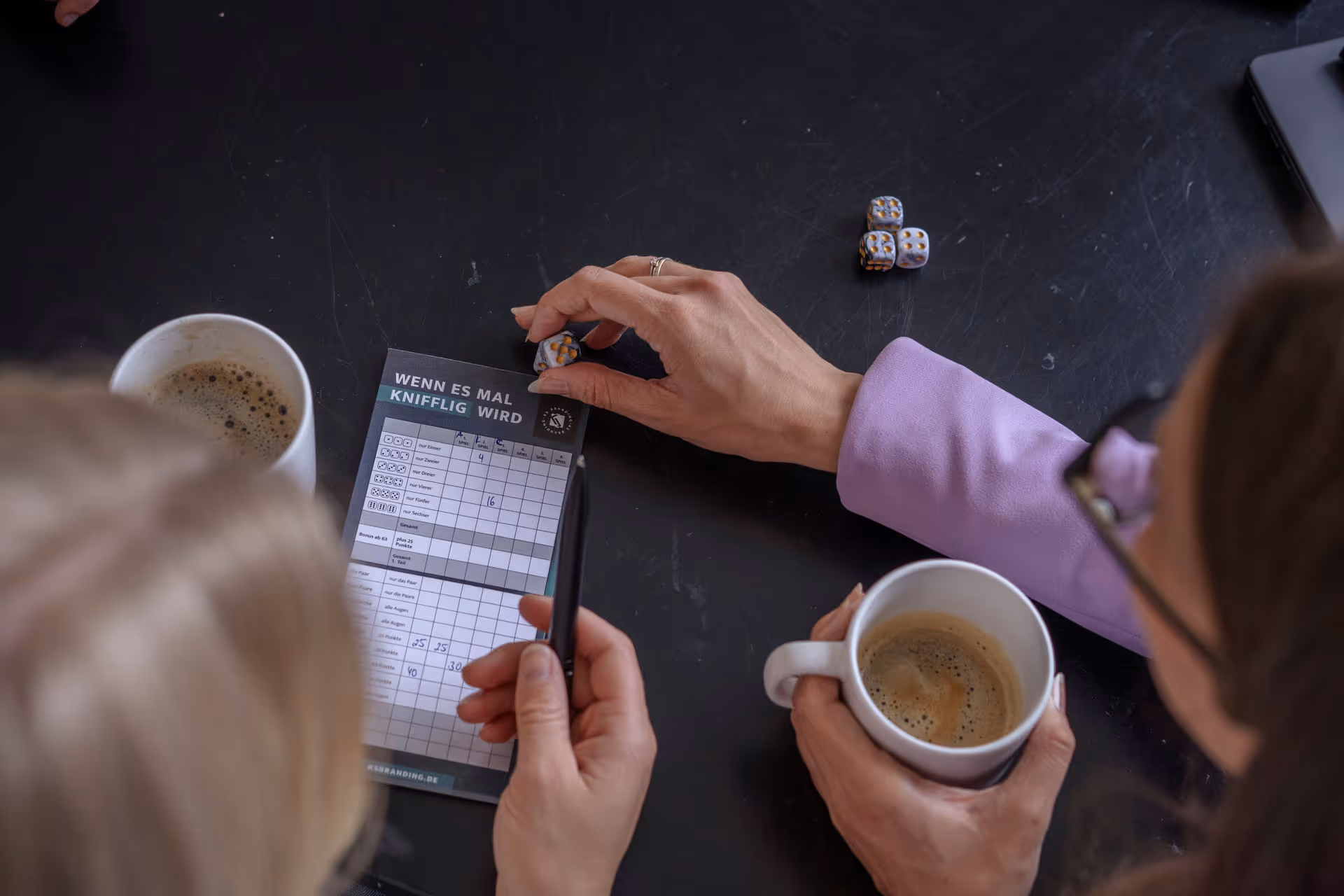 Zwei Personen spielen ein Würfelspiel, eine hält eine Tasse Kaffee, die andere markiert Punkte auf einem Spielzettel.