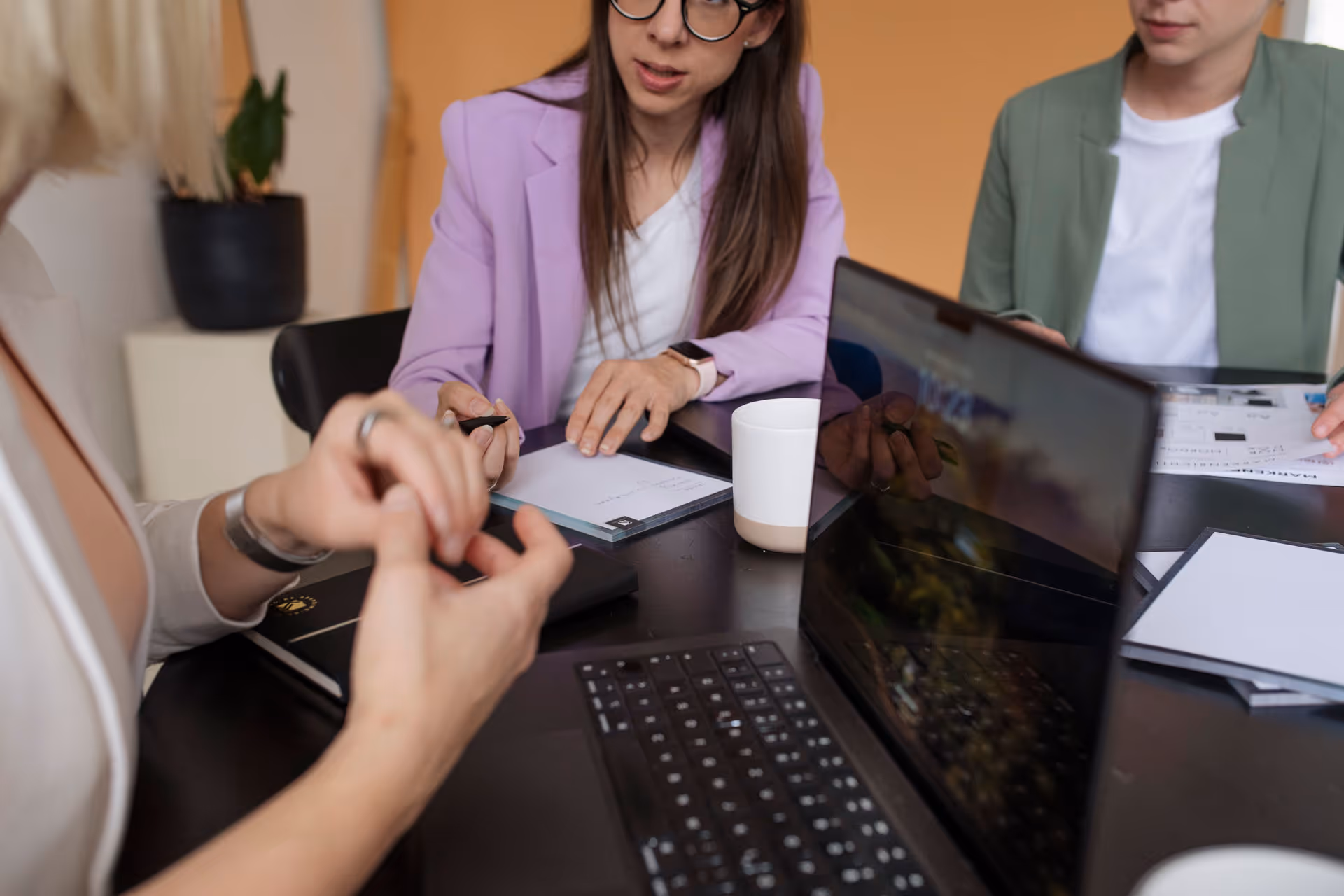 Drei Personen bei einer Besprechung am Tisch mit Laptop, Notizblöcken und einer Tasse.