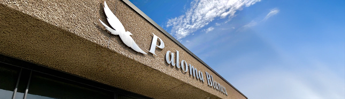 Building sign reading ‘Paloma Blanca’ with a white dove logo against a blue sky and scattered clouds.