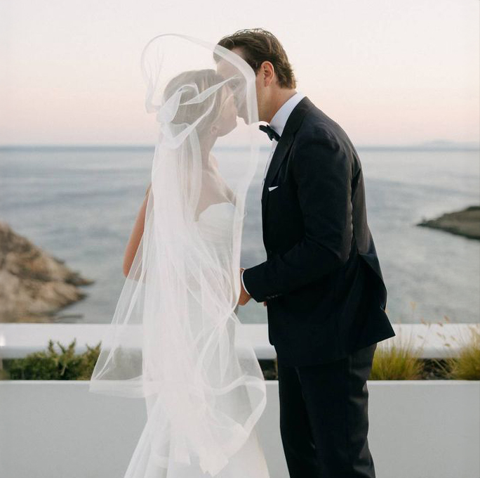 Bride and groom kiss outdoors by the ocean, with bride's veil blowing in the wind.