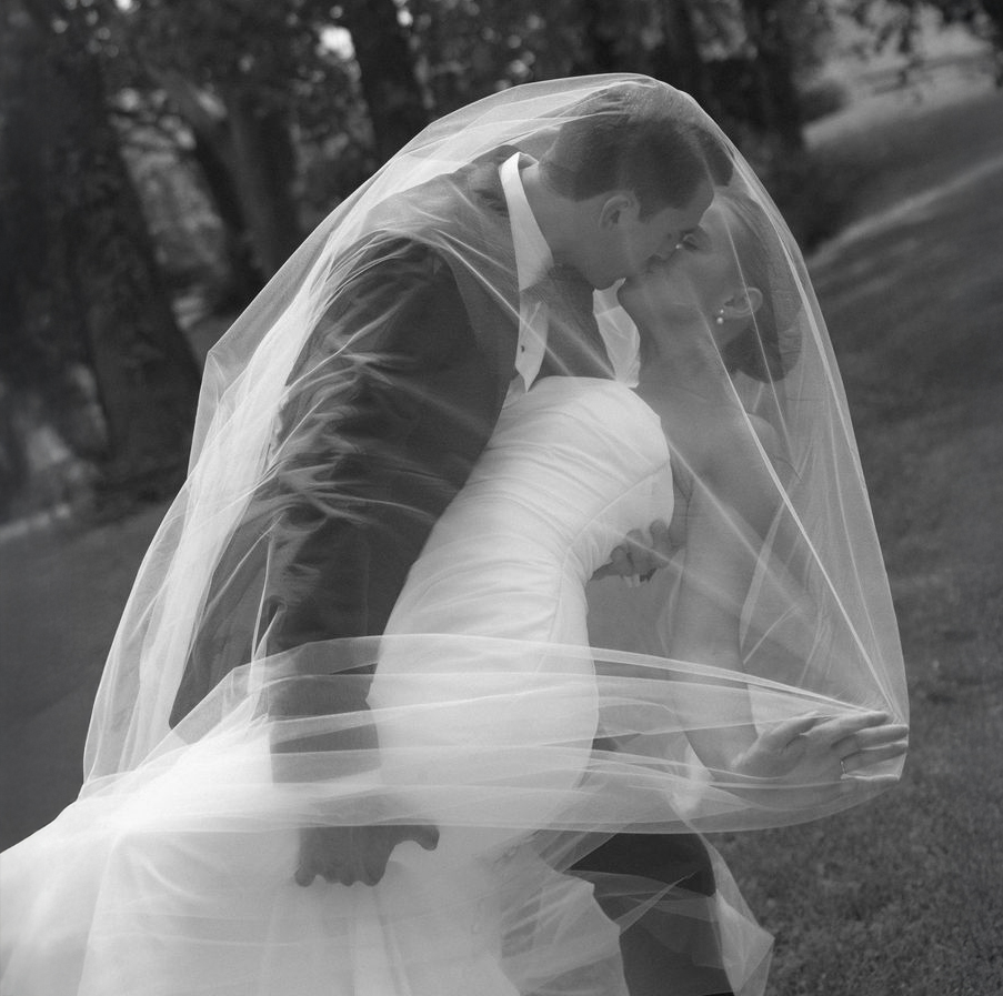 Black and white photo of a bride and groom kissing under the bride's veil outdoors.