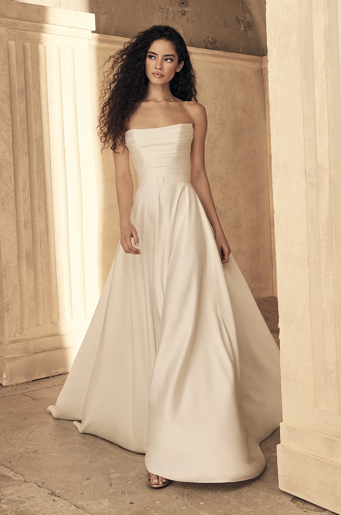 Strapless A-line gown with straight neckline, structured pleated bodice, slim satin belt, and flowing full skirt.