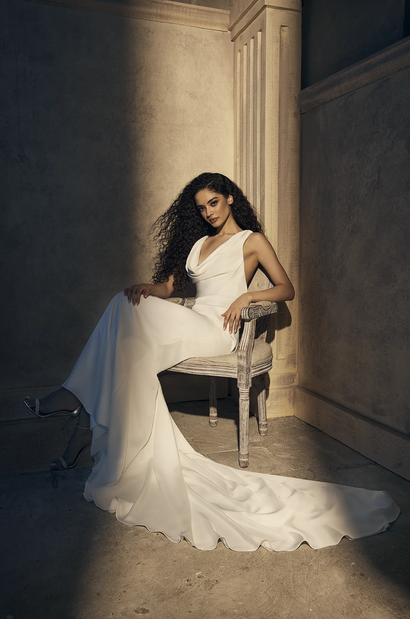 Wedding dress close-up showing draped cowl neckline, fitted satin bodice detail and flowing gown. Model sitting sideways.