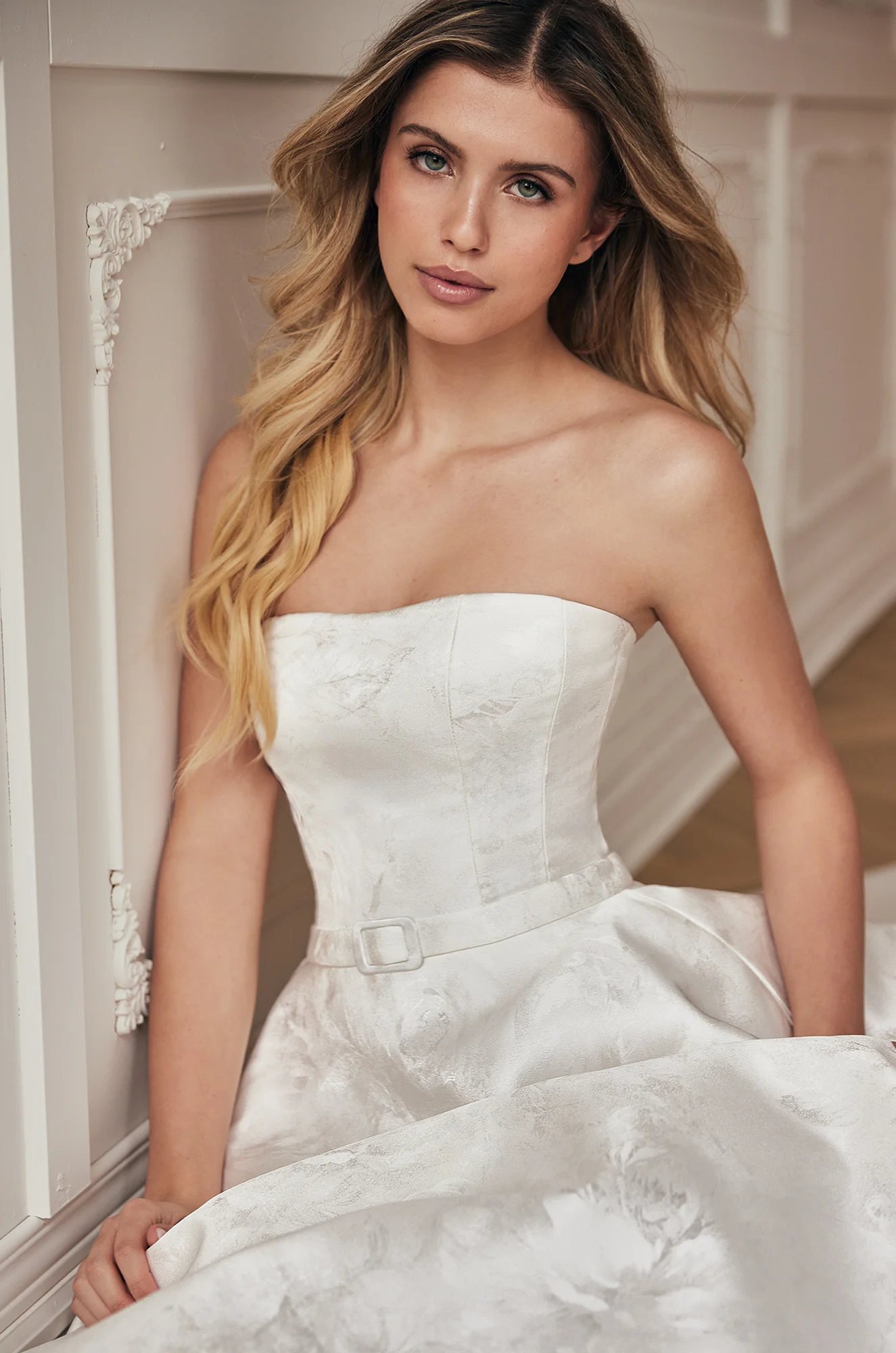 Close up view of strapless Jacquard wedding dress with straight across neckline and band at waist with buckle, in a ballroom.