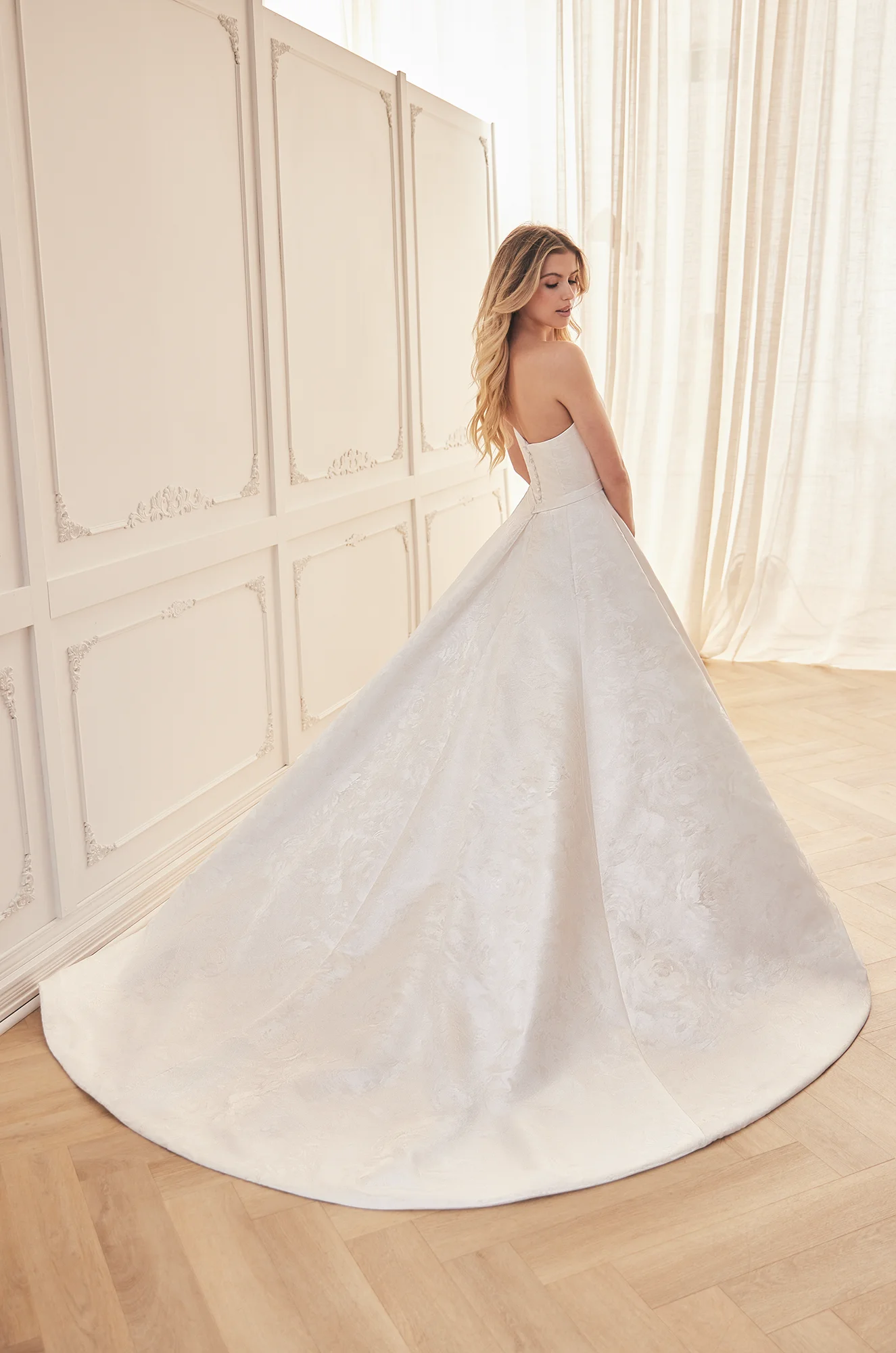 Back view of strapless Jacquard wedding dress with open back, buttons on back of bodice to waist, and ball gown skirt, in a ballroom.