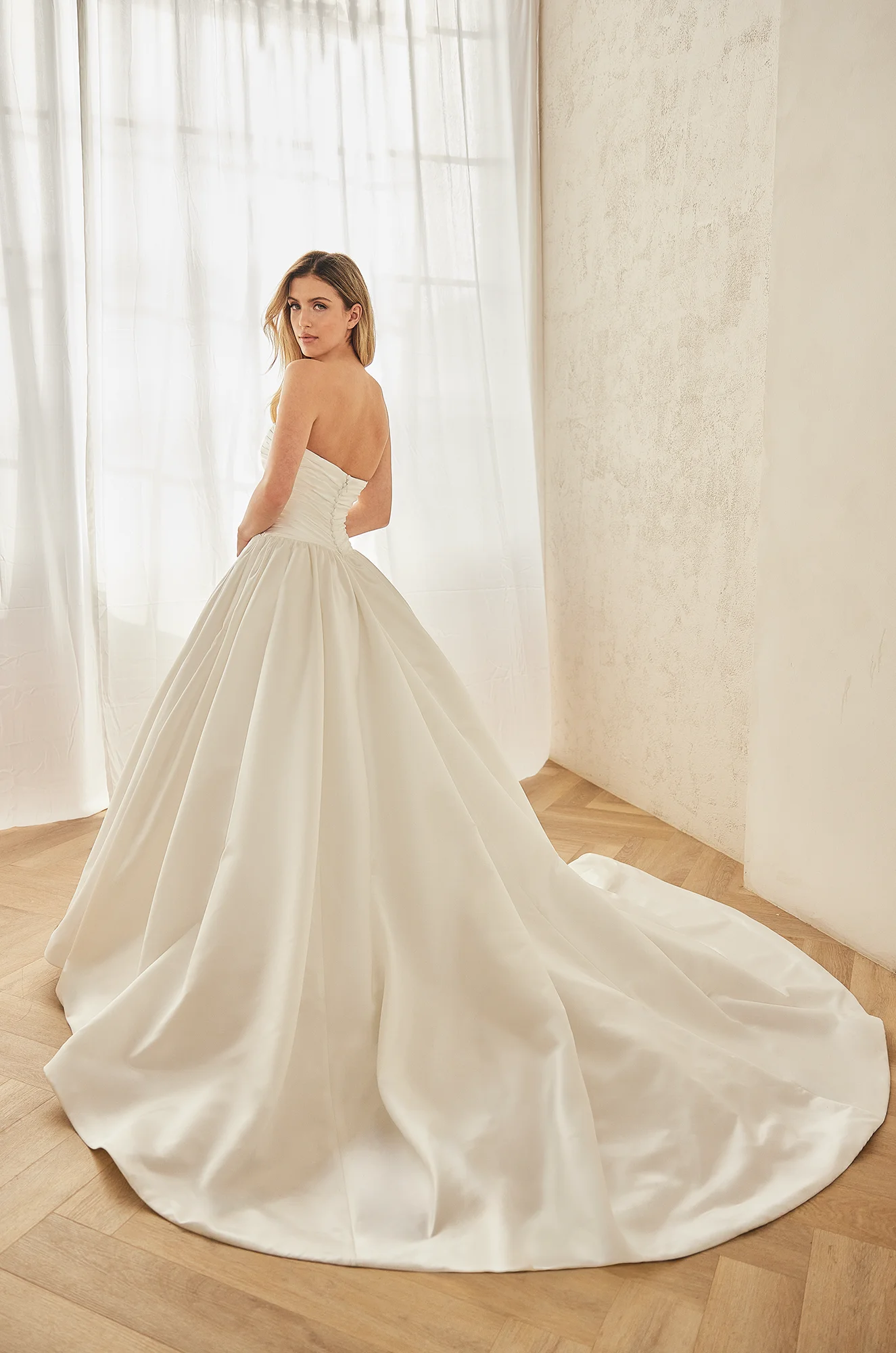 Back view of a strapless Satin wedding dress with a ruched drop waist bodice, buttons on the back of the bodice to waist, a ball gown skirt with gathers at the waist, in a ballroom.