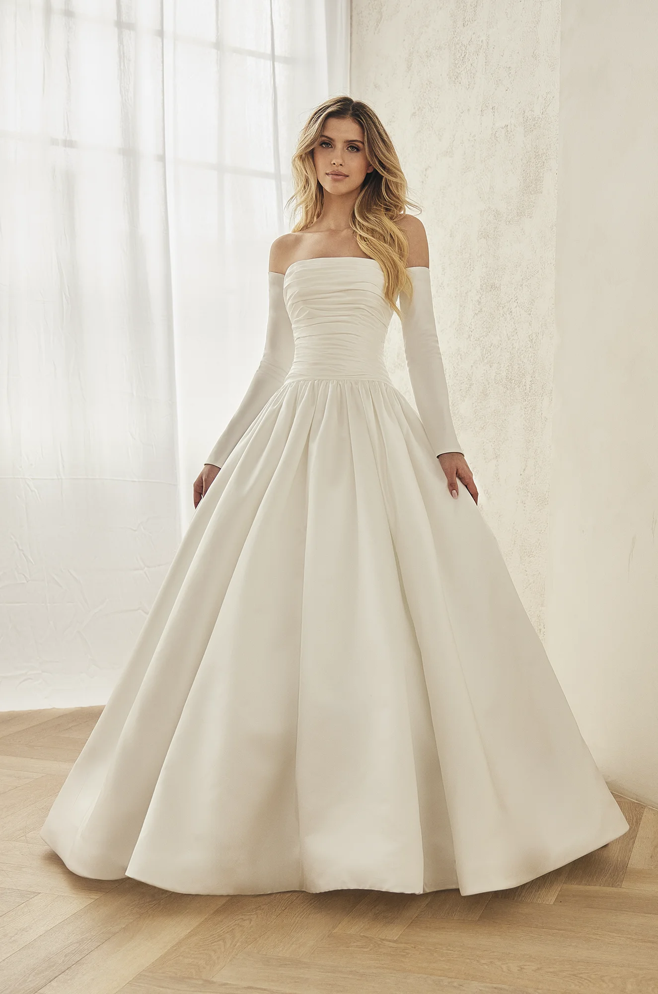Front view of a strapless Satin wedding dress with a ruched drop waist bodice, separate long Satin sleeves, a ball gown skirt with gathers at the waist, in front of a window.