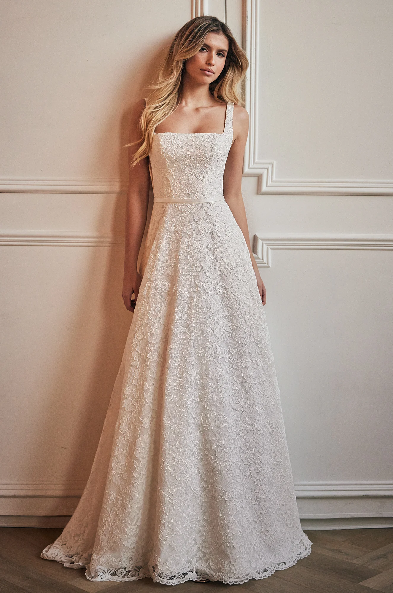 Front view of a sleeveless lace wedding dress with a square neckline and cut outs on the side of the bodice, a fabric band at the waist, with a lace A-line skirt, in a ballroom.