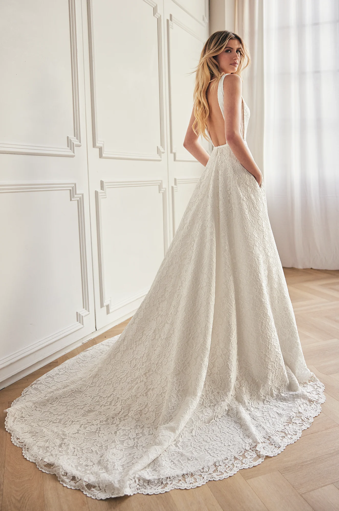 Back view of a sleeveless lace wedding dress with an open back, and a lace A-line skirt with pockets and lace scallop on the hem, in a ballroom.