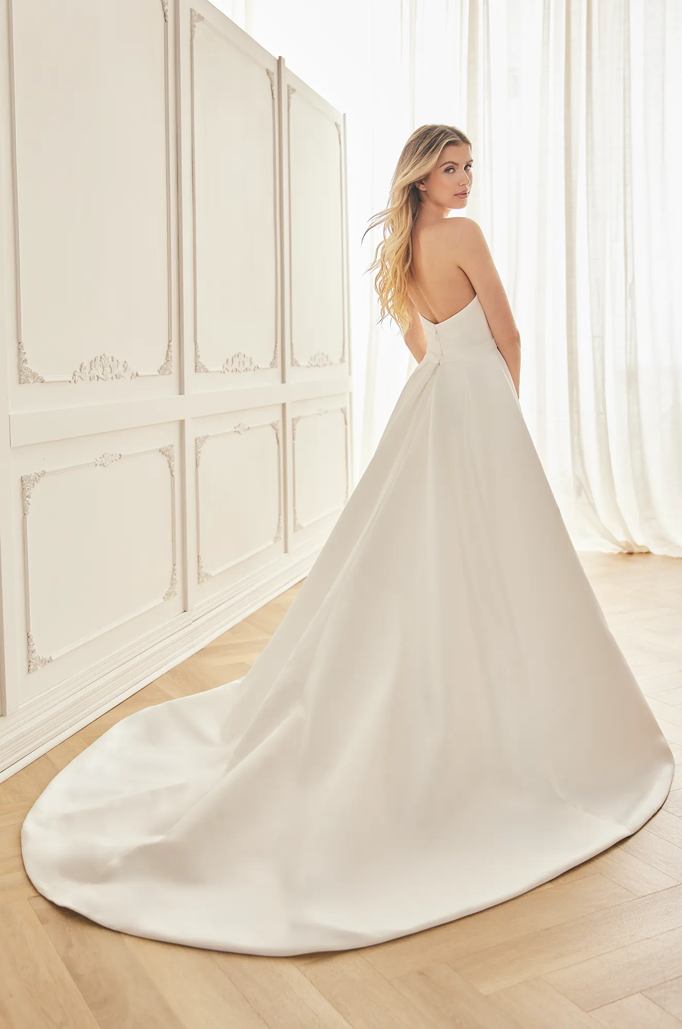 Back view of a strapless Satin Faille wedding dress with an open back, a V-shaped back neckline and a full skirt with pleats at waist, in a ballroom.