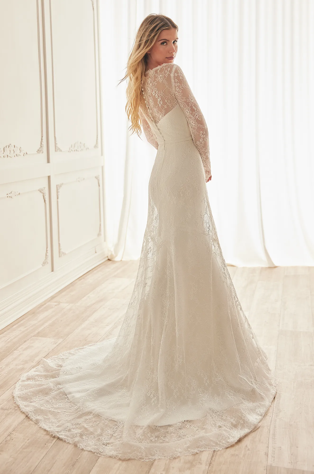 Back view of a strapless Crepe wedding dress with lace overlay that has long sleeves and full lace back with buttons down entire back to waist, with A-line lace overlay over fit and flare Crepe skirt, in a ballroom.