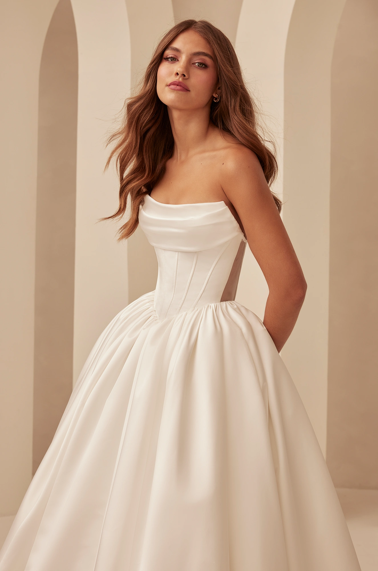 Close up view of a strapless Duchesse Satin wedding with a draped neckline, boning detail on bodice, a basque waist, and a gathered ball gown skirt, in front of pillars.