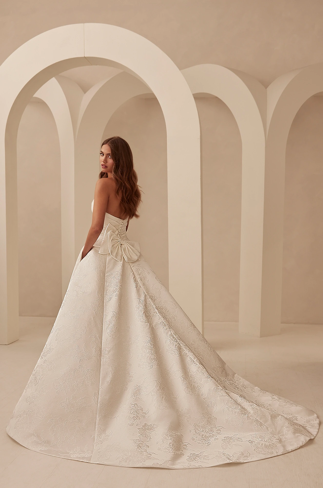 Back view of a strapless, floral print Jacquard wedding dress with buttons on back of bodice to waist, a large bow at the back of the waist, and an A-line skirt with pockets, in front of pillars.
