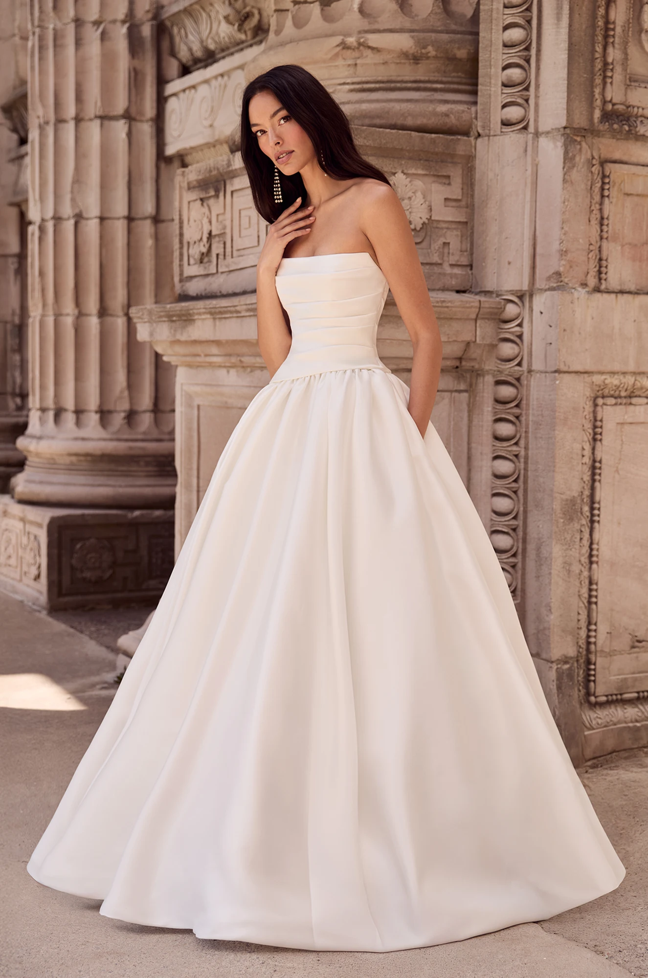 Strapless ball gown with pleated bodice, defined waist, and full gathered skirt, set against ornate stone architecture.