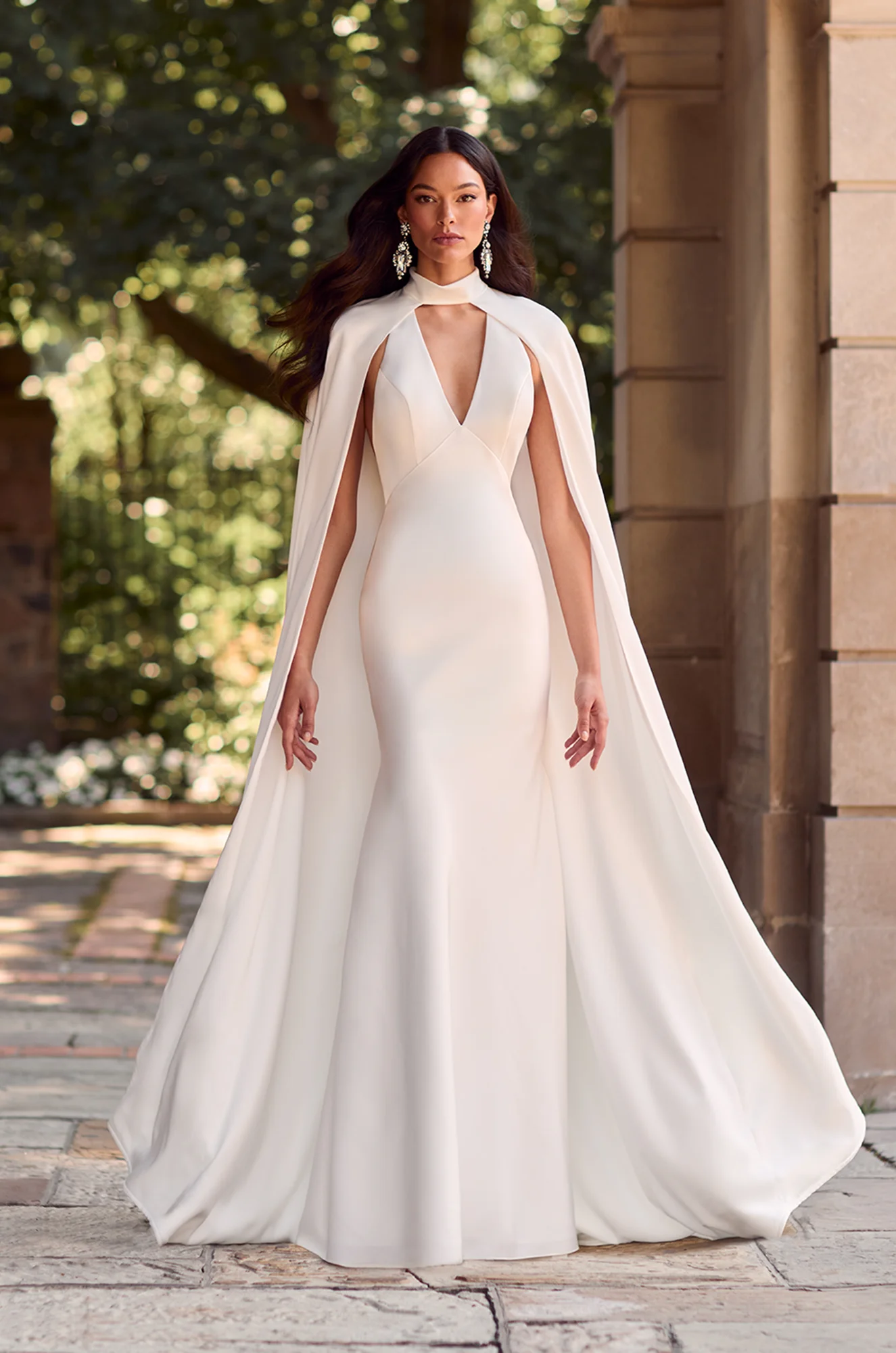 Fitted gown with deep V-neckline and floor-length cape, creating a sleek and dramatic modern bridal look.