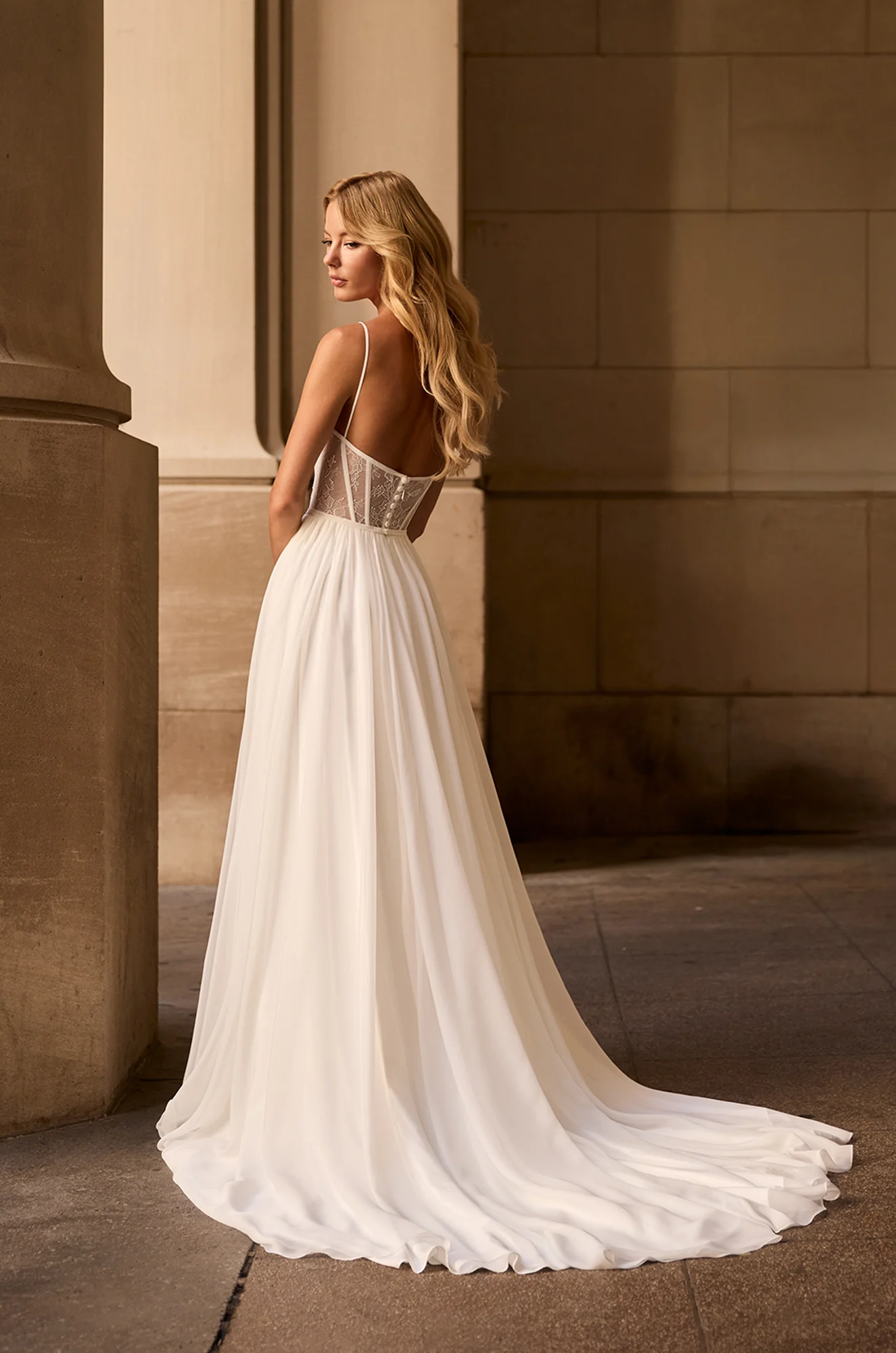 Back view of Chiffon wedding dress with sheer lace bodice, spaghetti straps, with exposed boning on back and buttons on back of bodice to waist, and a Chiffon A-line skirt, outside of a building.