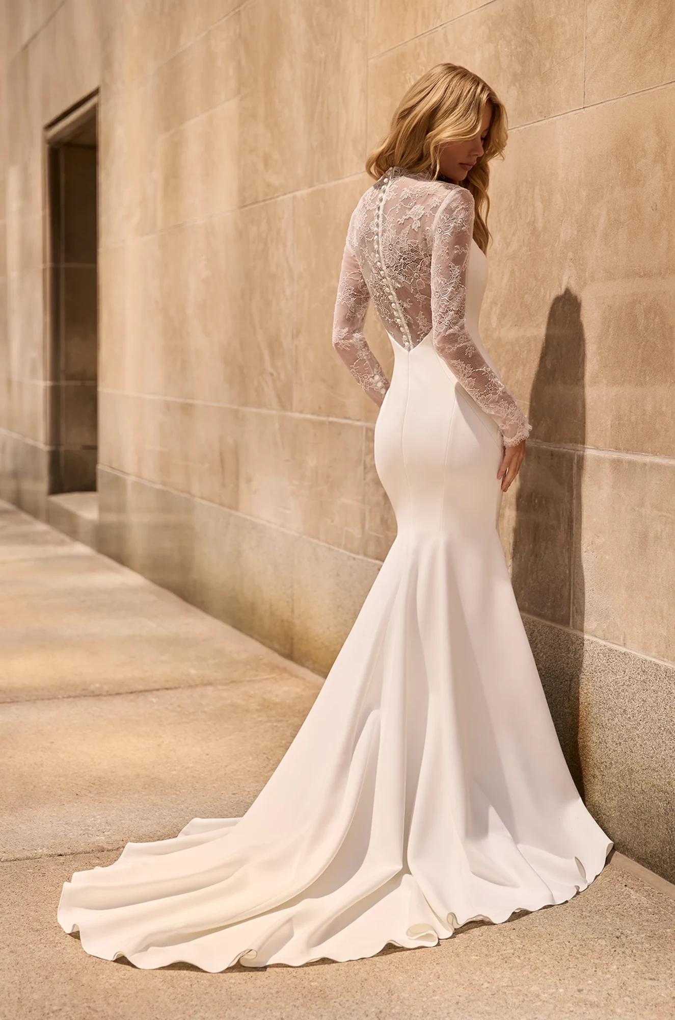 Back view of Crepe wedding dress with illusion lace on back and long lace sleeves, with buttons from back of neck to waist on sheer lace back and fit and flare Crepe skirt, outside of a building.