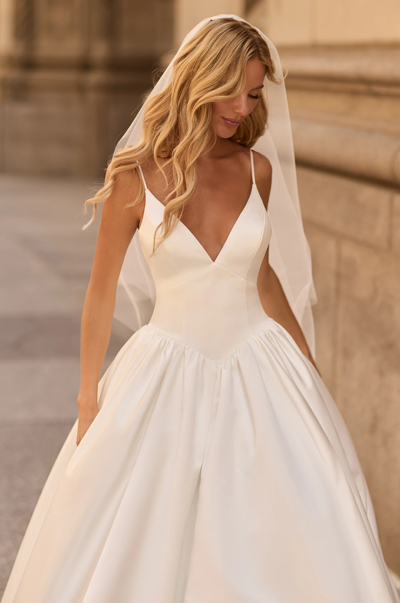 Close up view of Satin V-neckline wedding dress with spaghetti straps, basque waistline, and gathered ball gown skirt with pockets, outside of a building. Model is wearing a tulle veil.