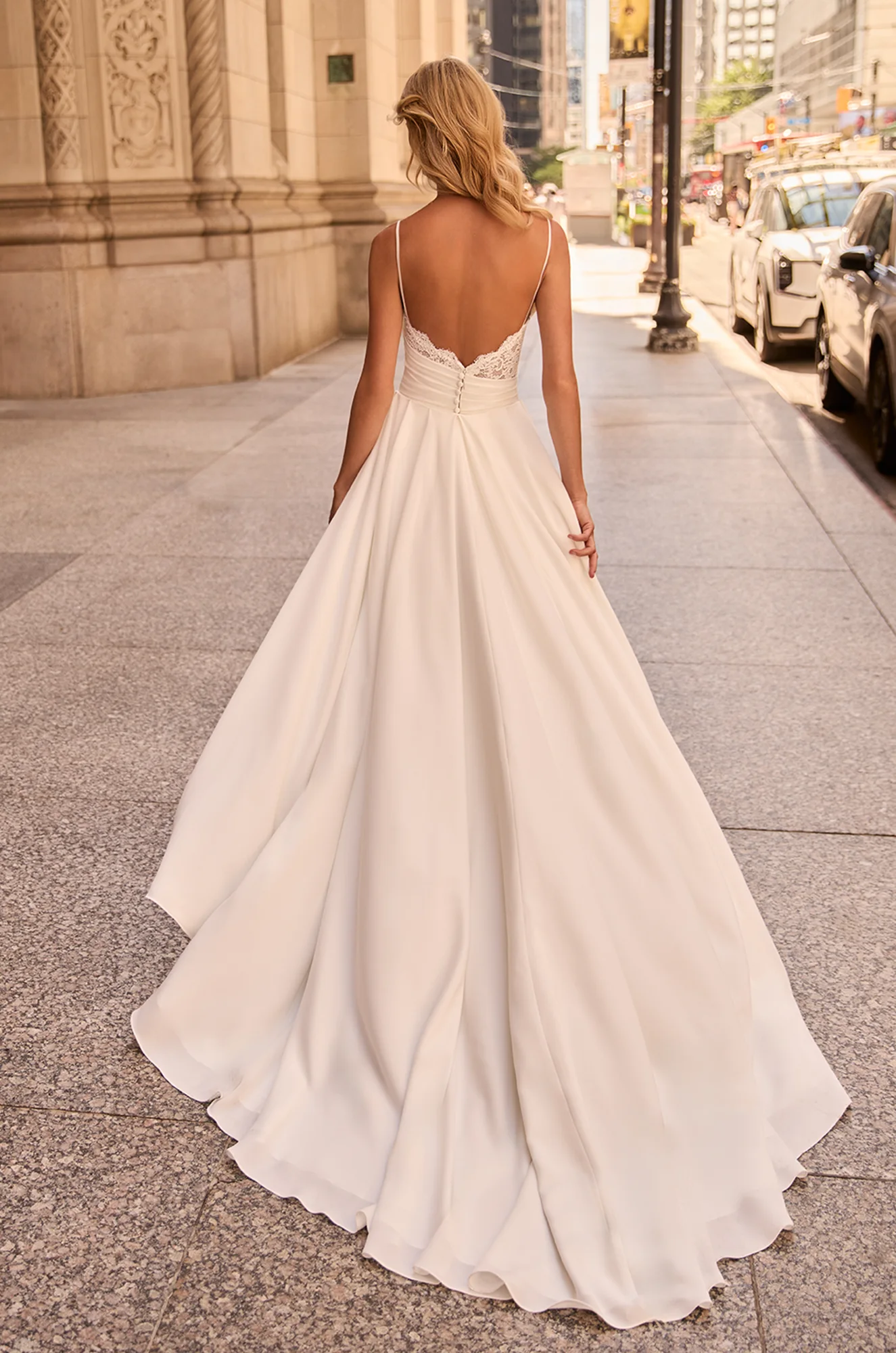 Back view of wedding dress with lace along back neckline, spaghetti straps, buttons on back of bodice, and removable overskirt attached at the waist of the dress, outside of a building.