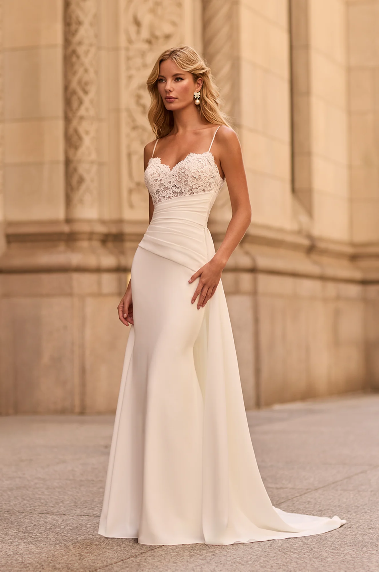 Front view of sweetheart neckline wedding dress with sheer lace bodice, spaghetti straps, asymmetric draped midriff, and fit and flare Crepe skirt, outside of a building. There is a Crepe overskirt attached at the waist of the dress.
