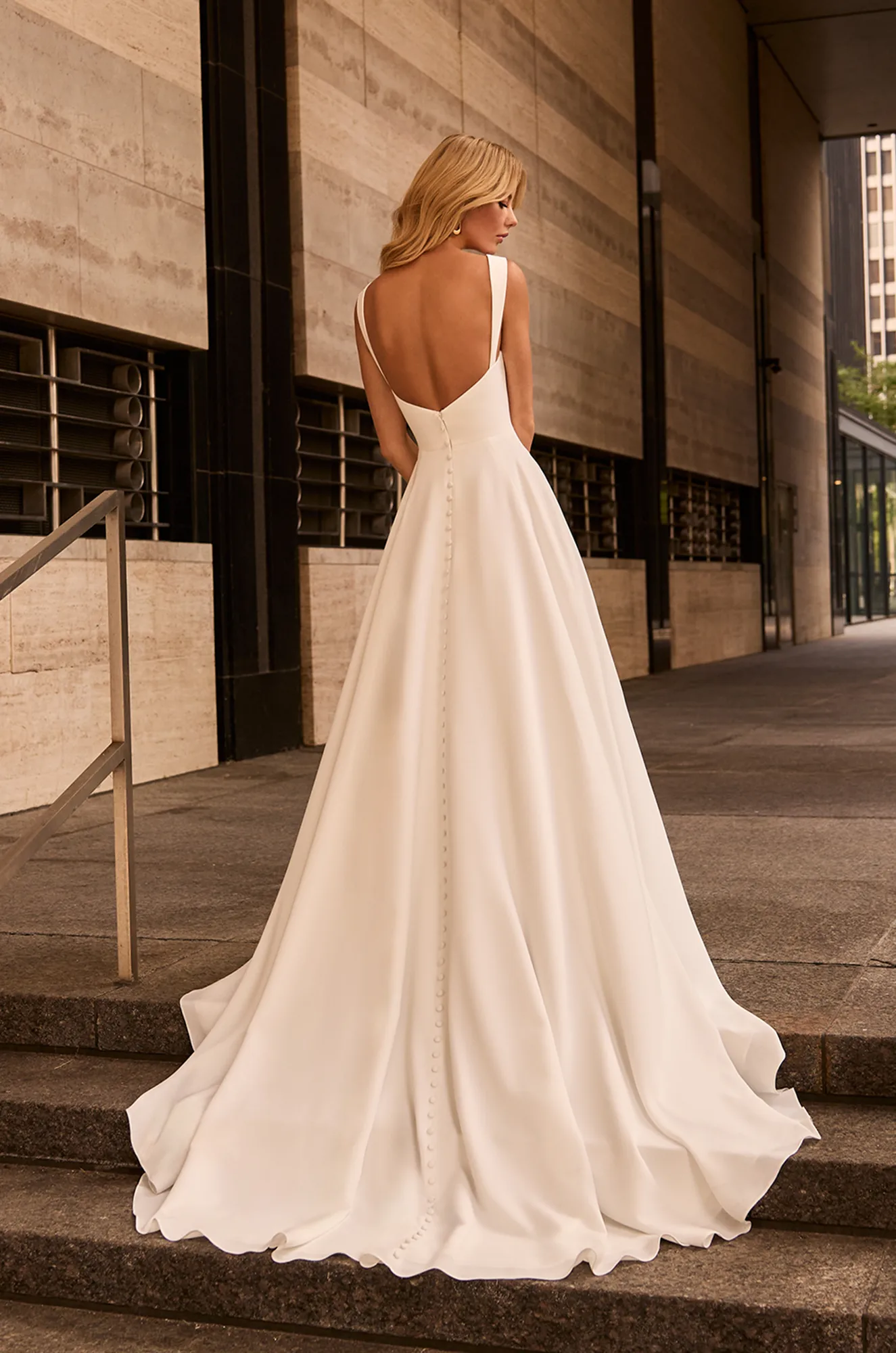 Back view of sleeveless Crepe wedding dress with slight V-back neckline, an A-line skirt, buttons down back of bodice and skirt, outside of a building.