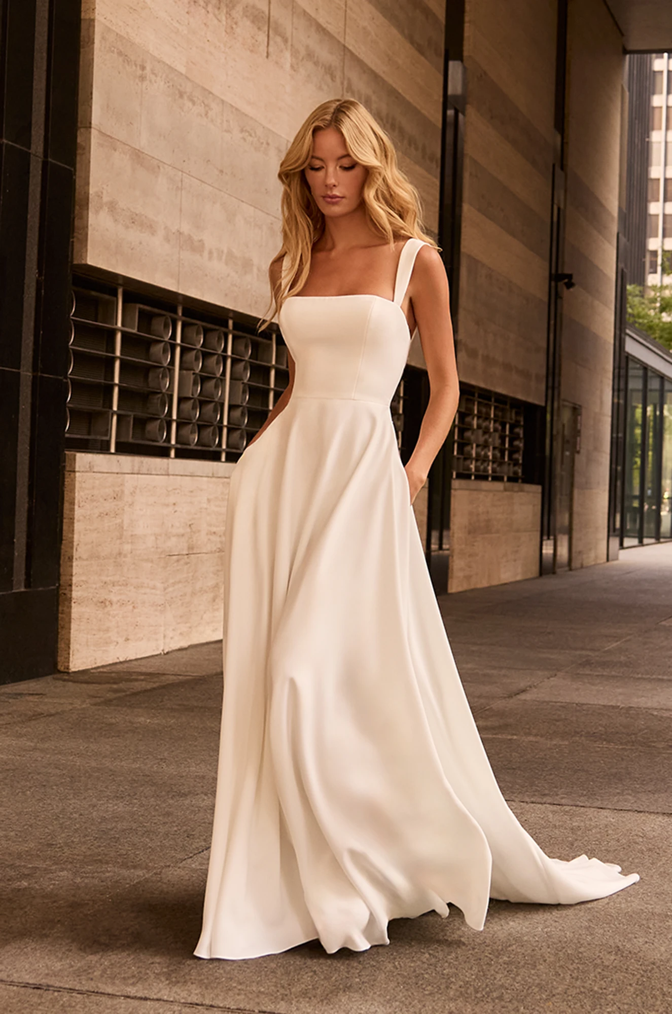 Front view of sleeveless Crepe wedding dress with square neckline and A-line skirt with pockets, outside of a building.