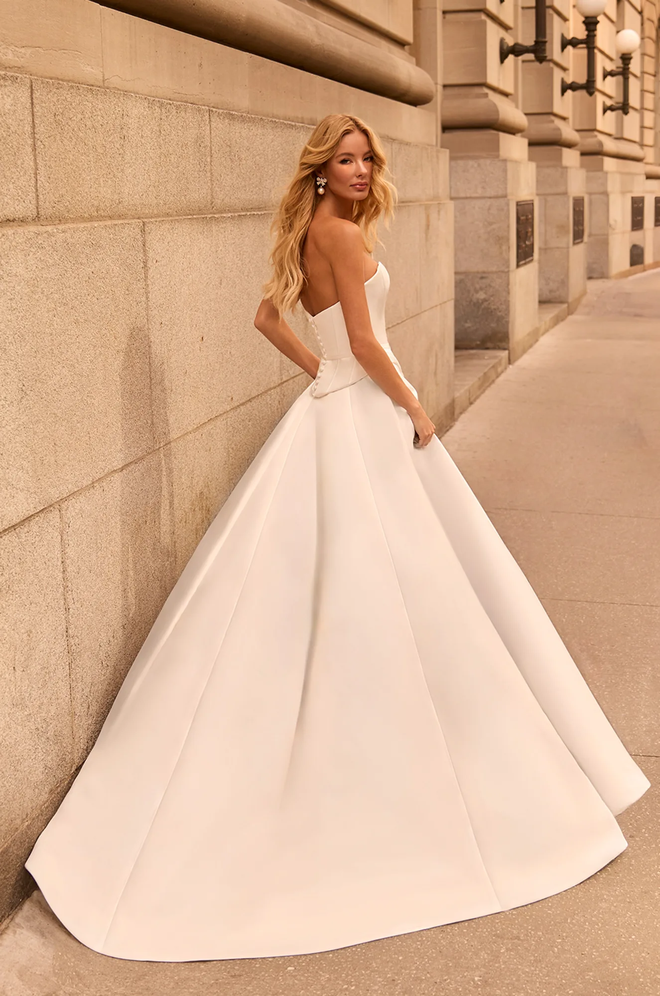 Back view of strapless two piece Satin wedding dress with open back,  buttons down back of bodice, and pleat on back of A-line skirt, outside of a building.
