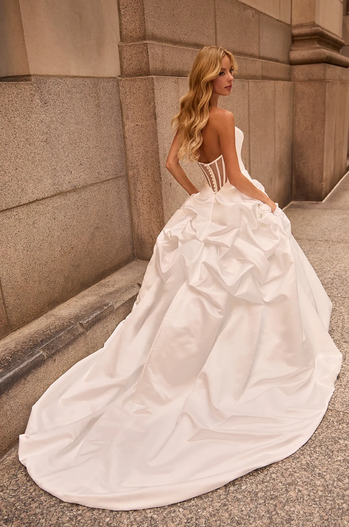 Back view of strapless Satin wedding dress with exposed boning on back of bodice and buttons down back to waist, and full pickup skirt, outside of a building.