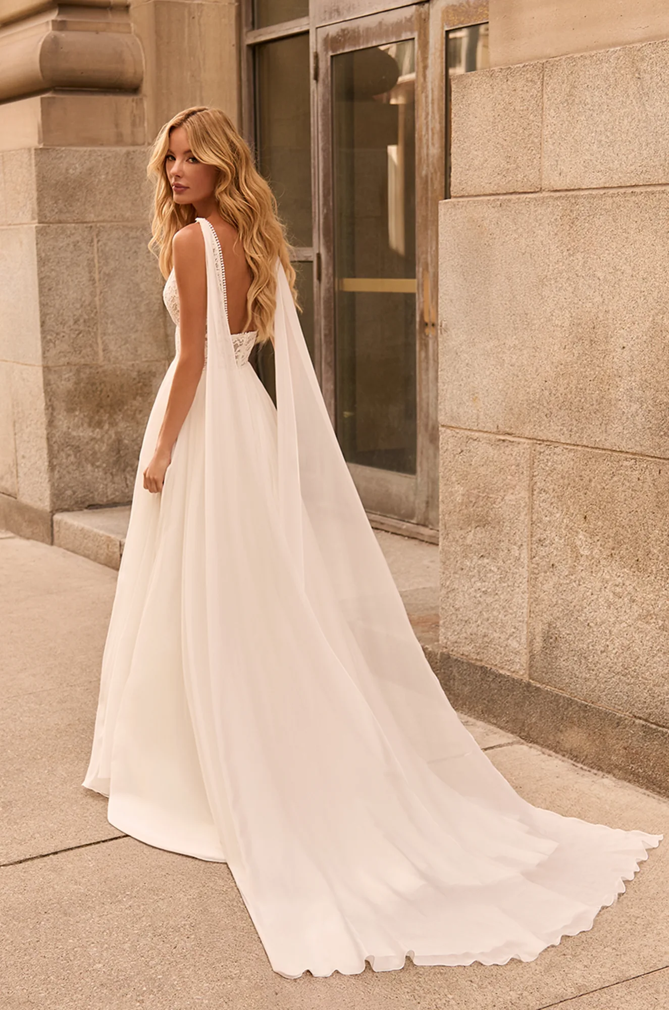 Back view of wedding dress with shoulder wings attached to sleeveless sheer lace bodice with Chiffon A-line skirt, outside of a building.