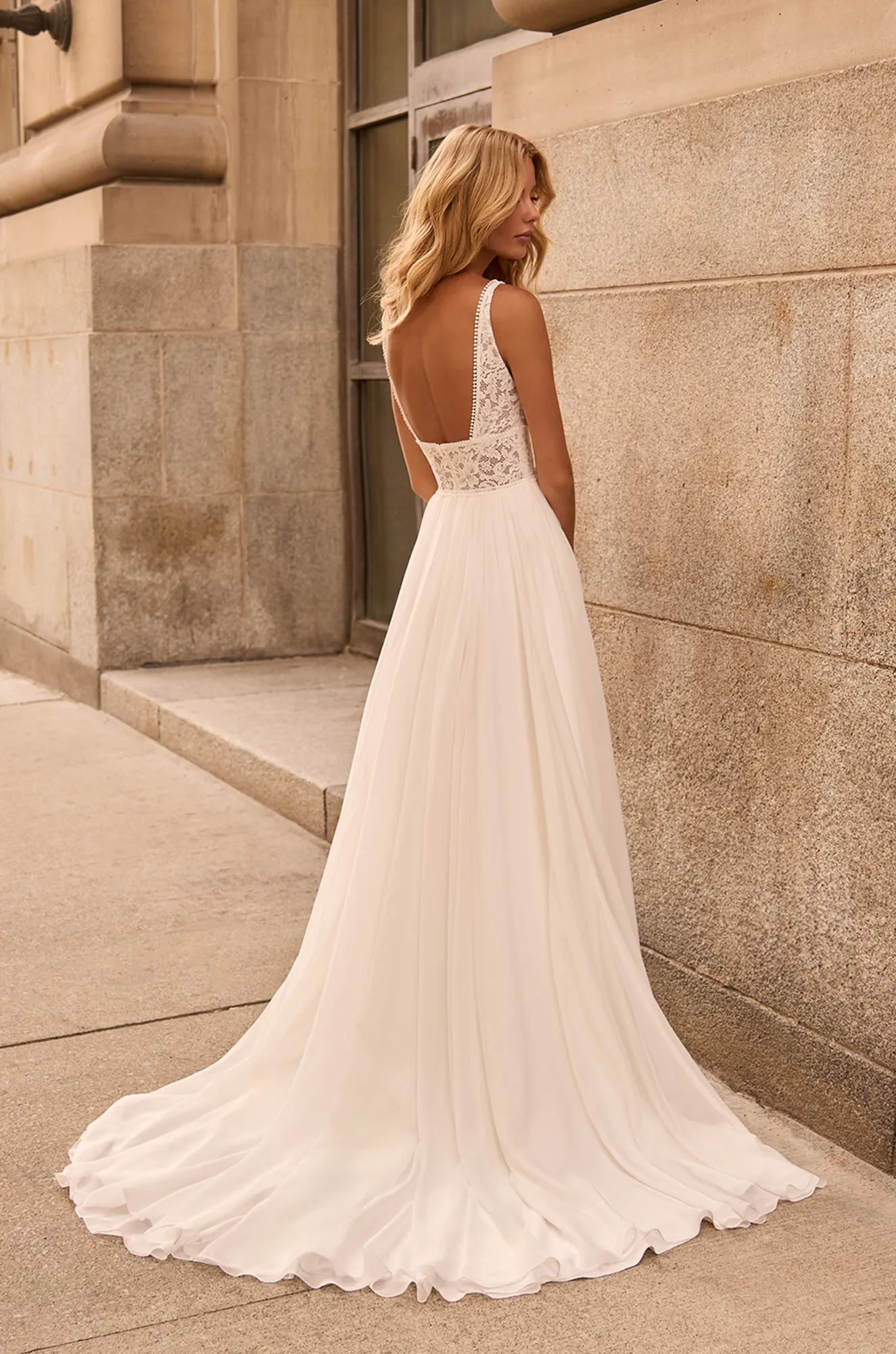 Back view of wedding dress with sleeveless sheer lace bodice and gathered Chiffon A-line skirt, outside of a building.