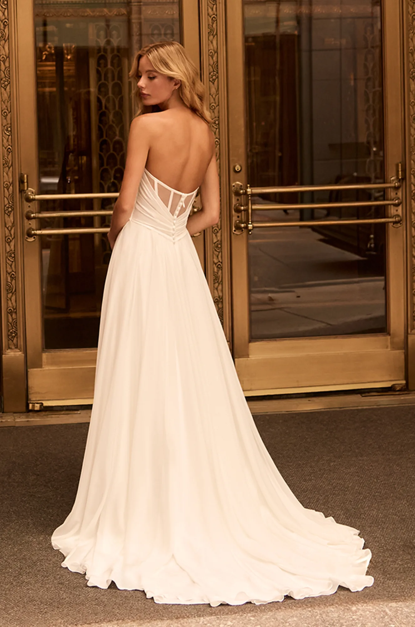 Back view of strapless Chiffon wedding dress with exposed boning on back, buttons on back to waist and A-line skirt, in front of glass doors.