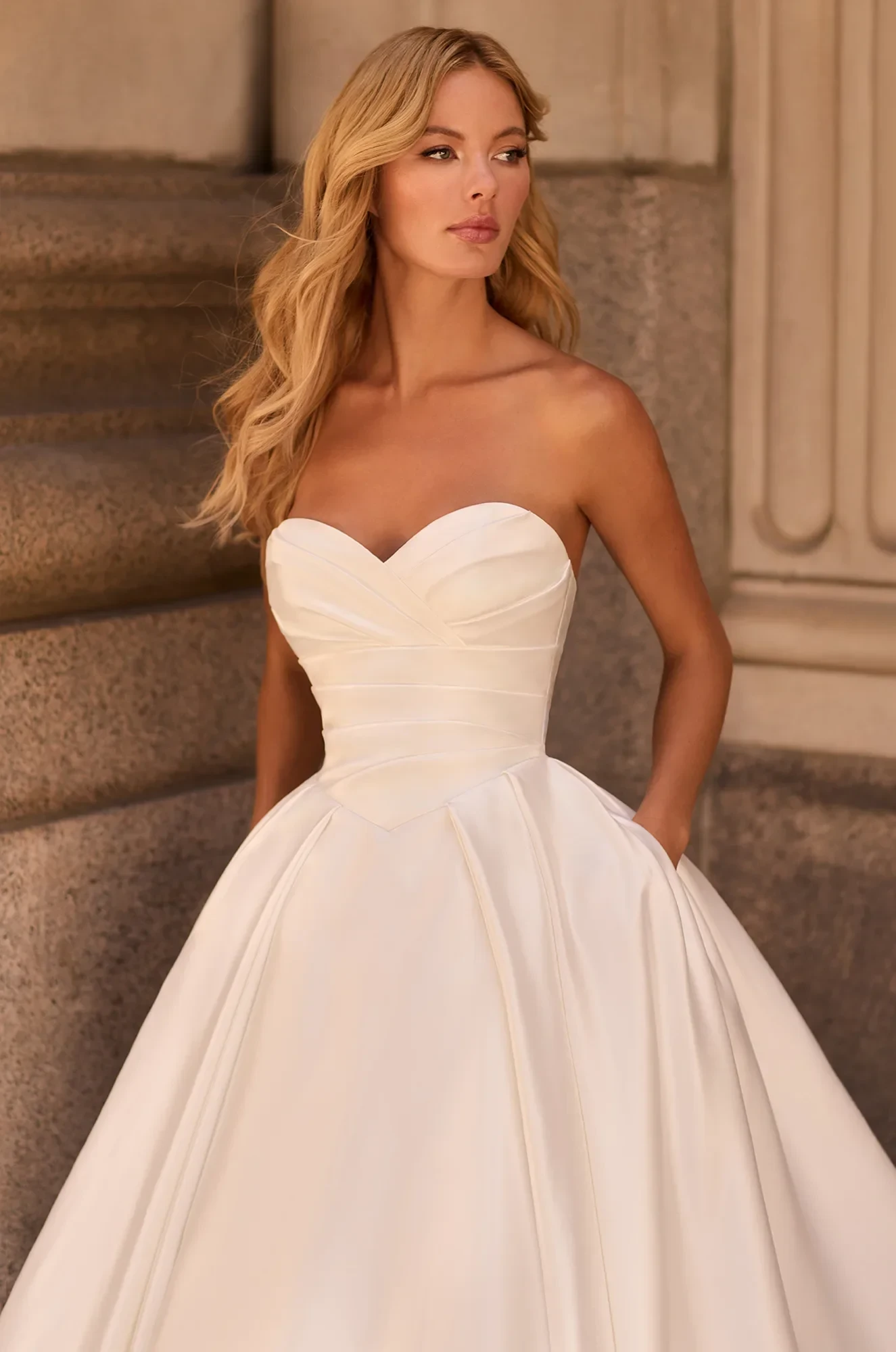 Close up view of strapless Satin ball gown wedding dress with pleated sweetheart neckline, basque waistline, and pockets, outside of a building.