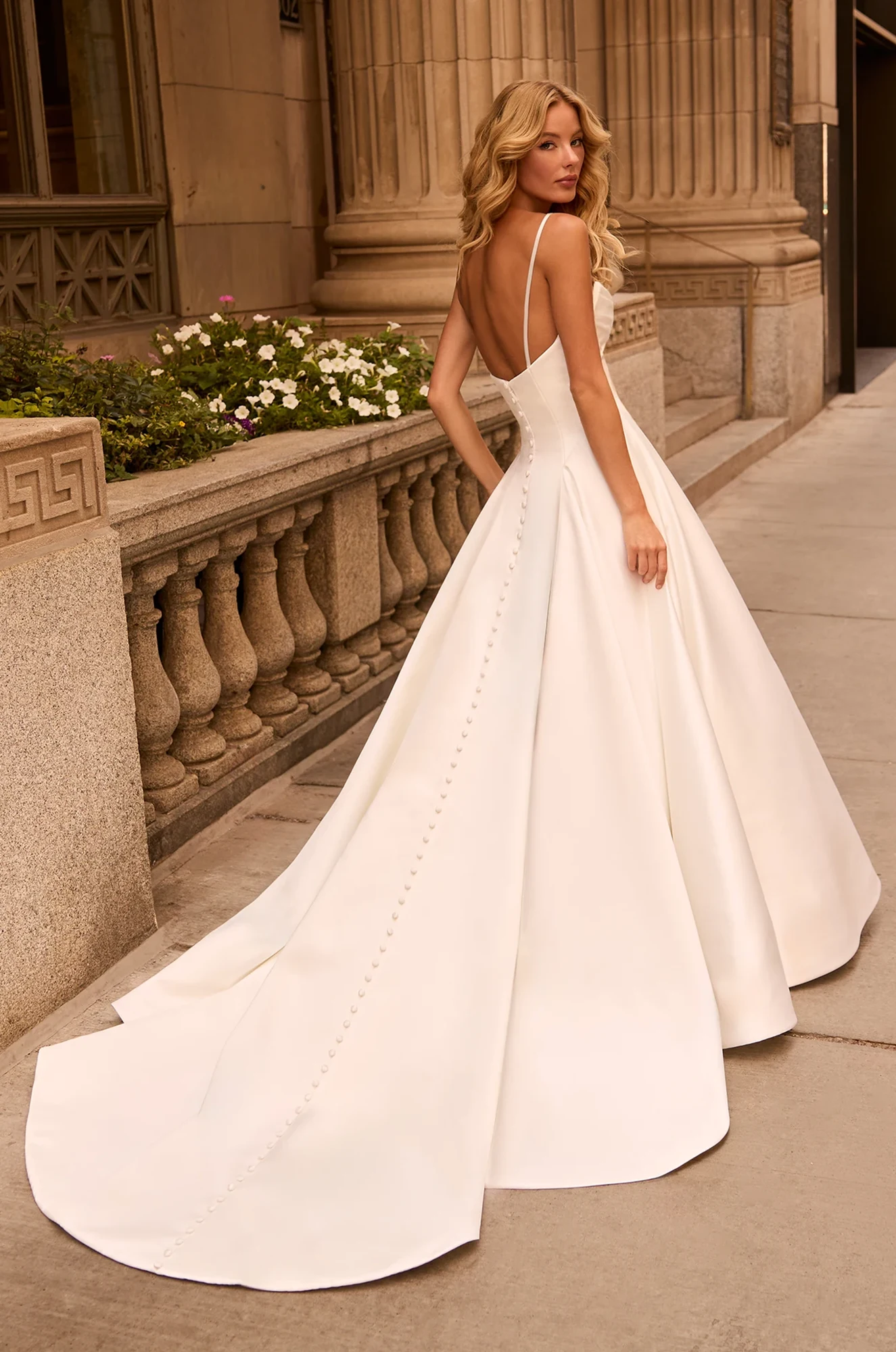 Back view of Satin A-line wedding dress with buttons down back of dress and a long train, outside of a building.