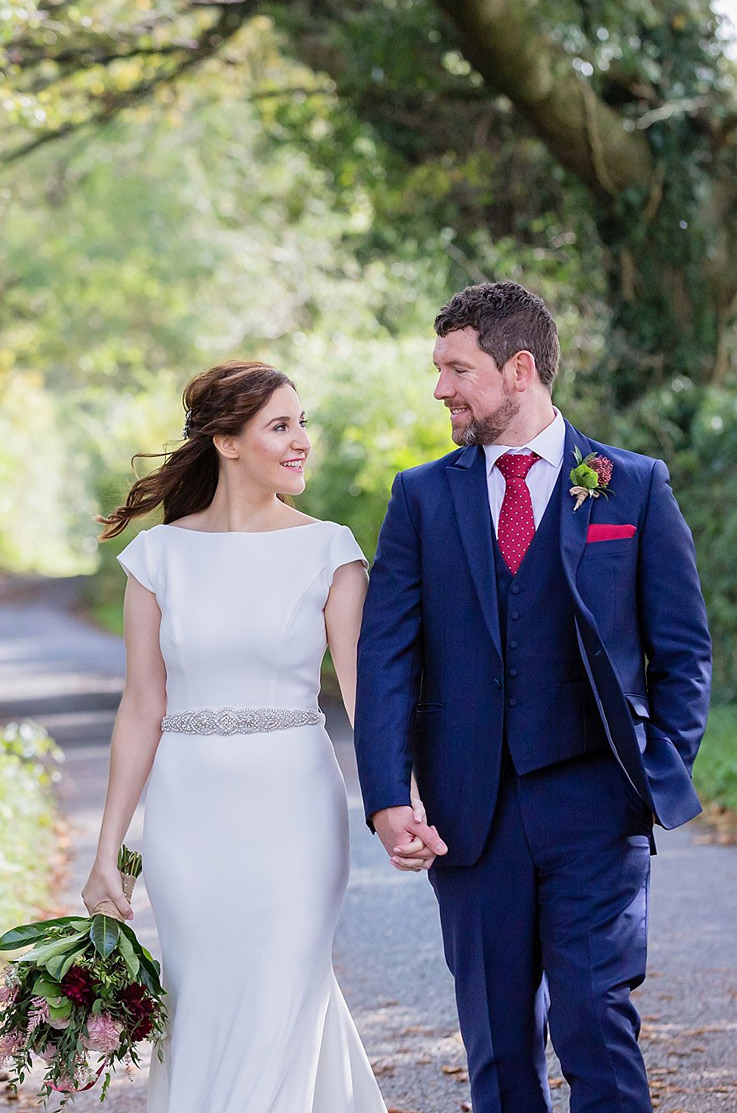 Bride in a sleek crepe wedding gown with cap sleeves, walks hand in hand with groom on a curved path, holding a bouquet.