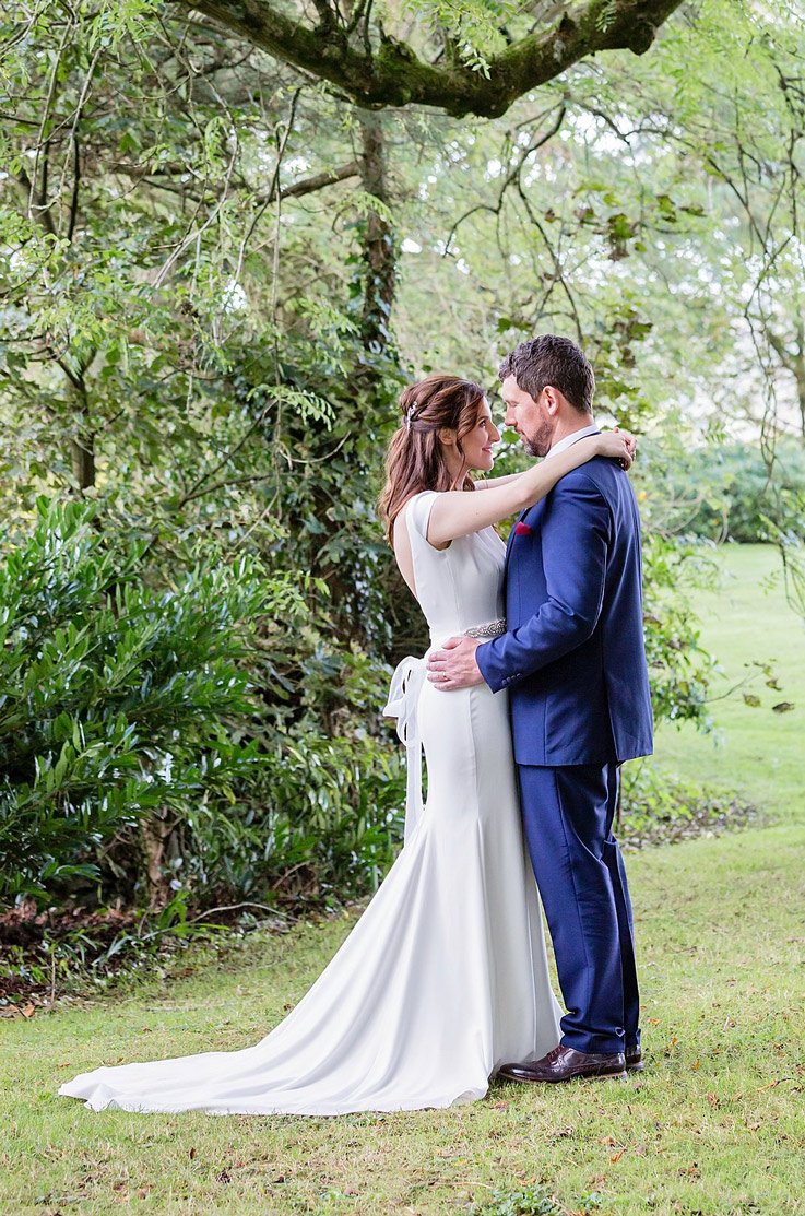 Bride in a cap sleeve crepe wedding gown with a beaded belt and tie, faces groom in a blue suit in an embrace, under a tree.