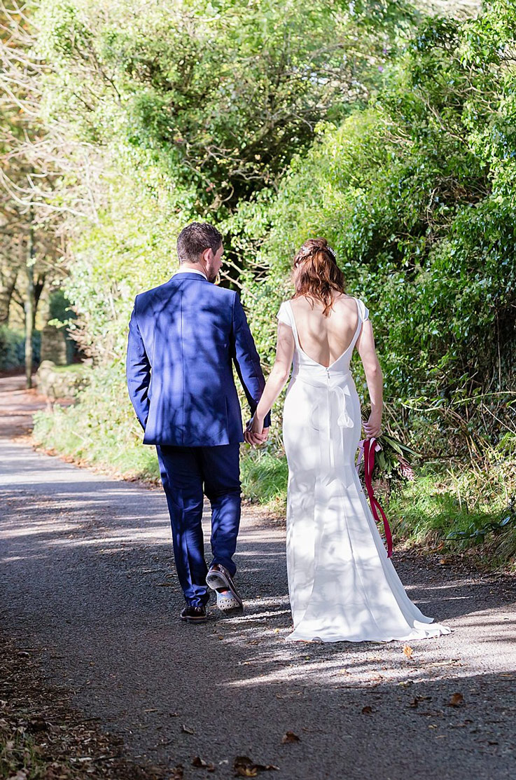 Bride in a crepe wedding dress with a boat neckline and beaded belt, holds hands with groom smiling on a tree-lined path.