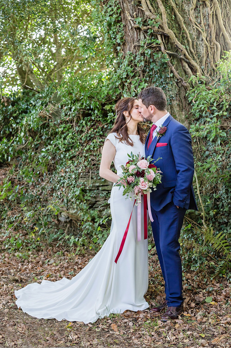 Bride in a fit and flare gown with cap sleeves and a chapel train, kisses the groom while holding a bouquet outdoors.