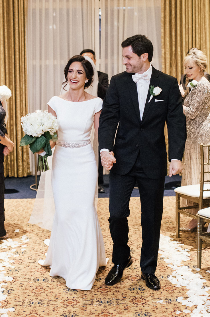 Bride in a fit and flare crepe gown with beaded belt detail, smiles and walks hand in hand with groom down the aisle.