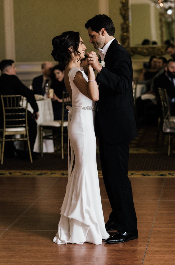  Bride in a cap sleeve crepe gown with a chapel train and beaded belt detail, dances with the groom in a black tuxedo. 